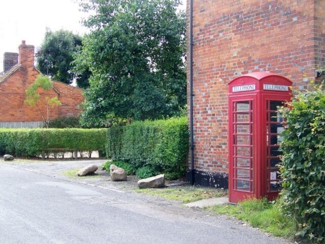 A red telephone booth next to a brick building with a hedge and a road.