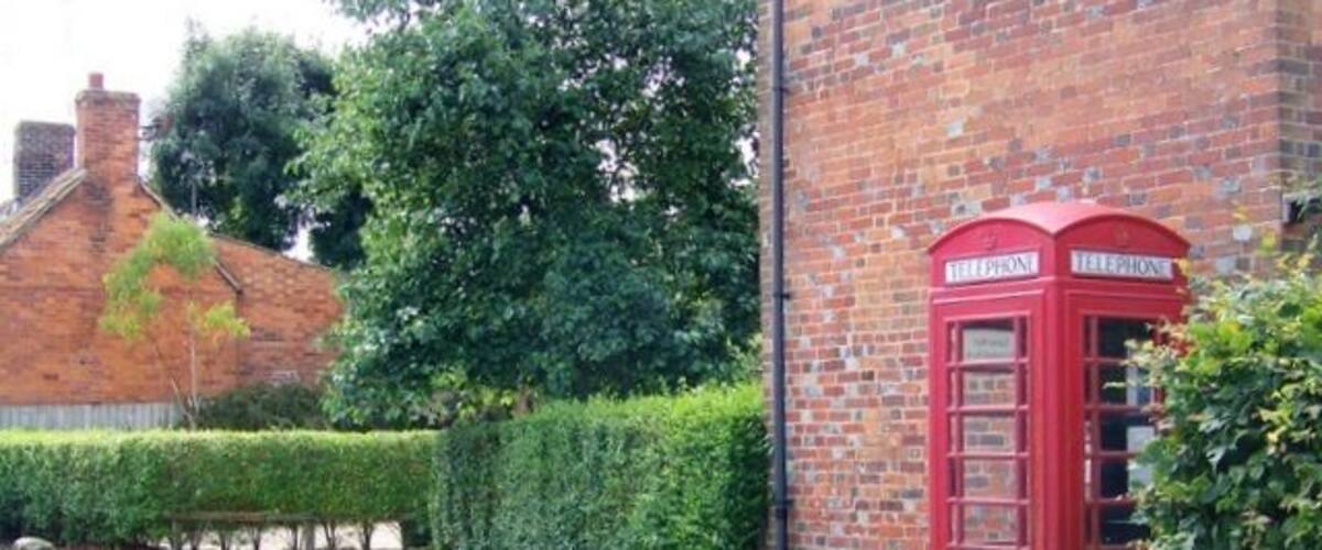A red telephone booth next to a brick building with a hedge and a road.