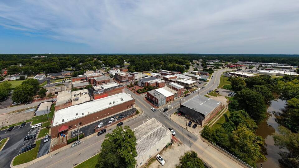 An aerial view of a town with a mix of commercial and residential buildings, a main road, and surrounding greenery.