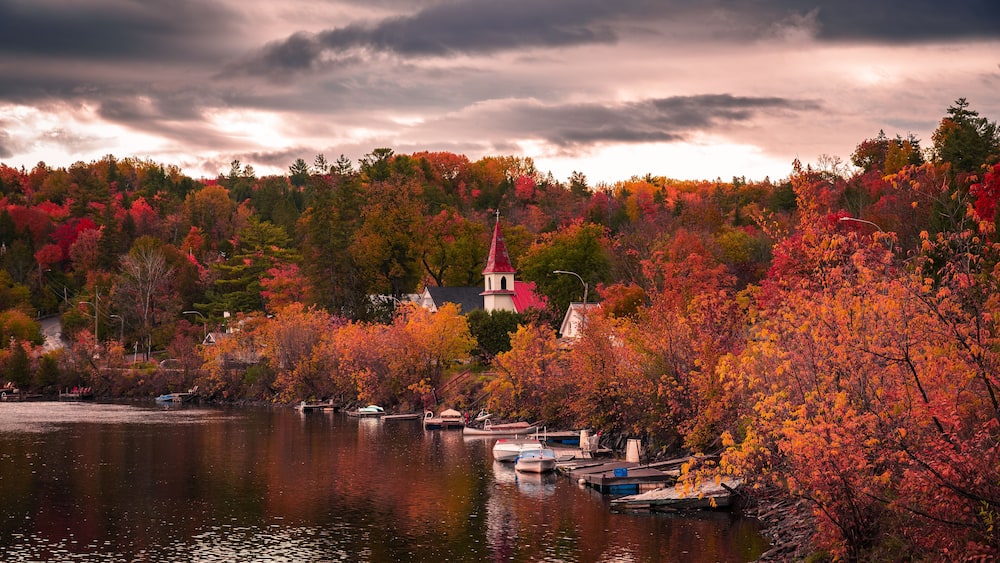 Autumn in picturesque village of Wakefield, boats on the Gatineau River, red church steeple, colourful foliage fall season, dramatic sky cloudscape, La Peche, Outaouais, Quebec, Canada, October 2022