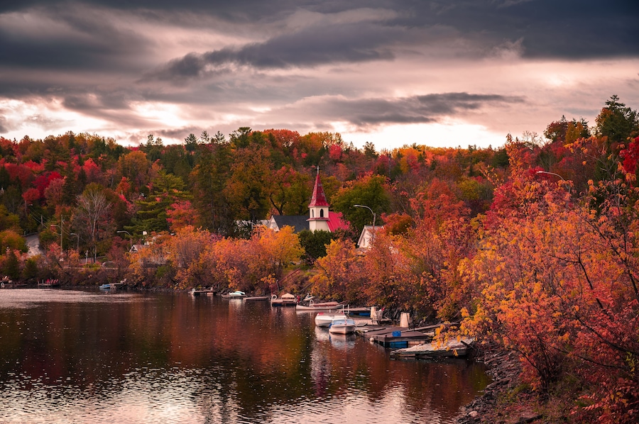 Autumn in picturesque village of Wakefield, boats on the Gatineau River, red church steeple, colourful foliage fall season, dramatic sky cloudscape, La Peche, Outaouais, Quebec, Canada, October 2022