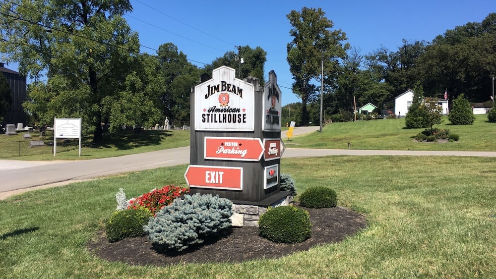 A Jim Beam American Stillhouse sign with an exit arrow, surrounded by landscaped bushes and flowers.