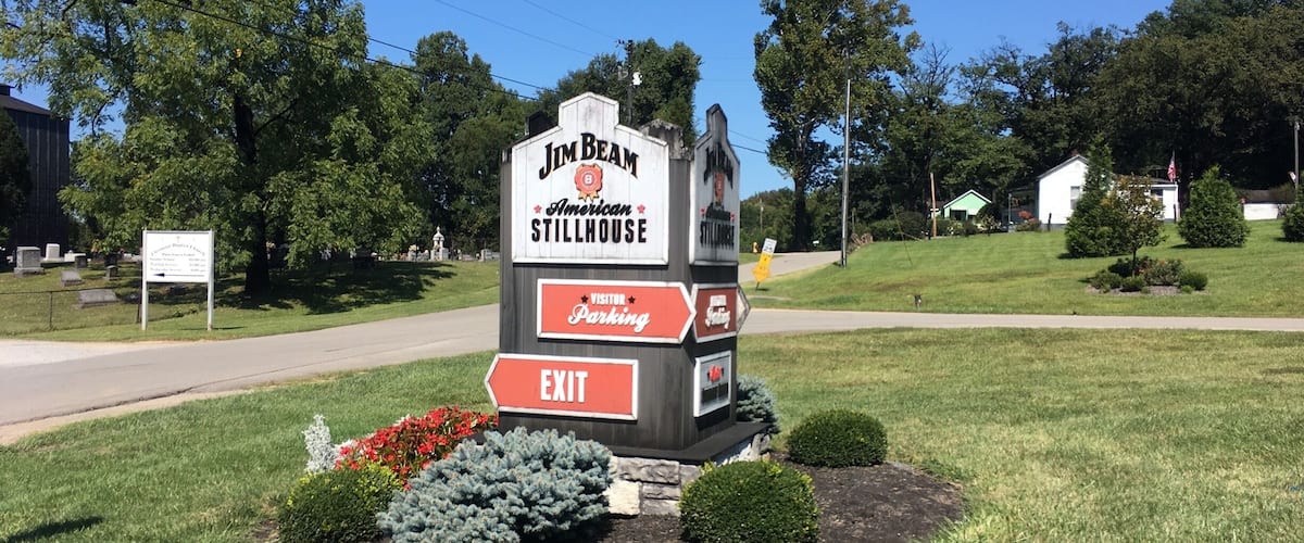 A Jim Beam American Stillhouse sign with an exit arrow, surrounded by landscaped bushes and flowers.