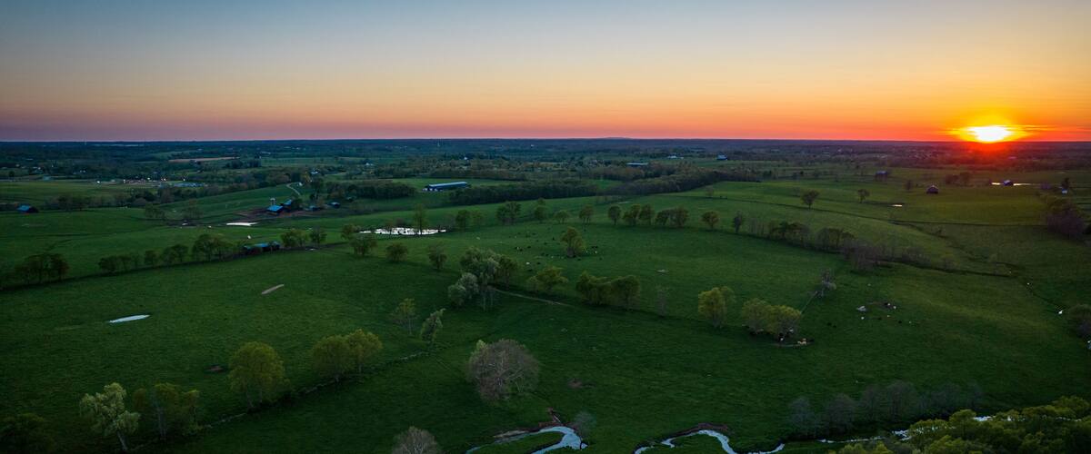 A sunset over a rural landscape with rolling green fields, a winding stream, and scattered trees.