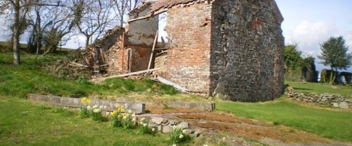 A dilapidated stone building with a chimney, surrounded by a grassy area with trees and a stone wall.
