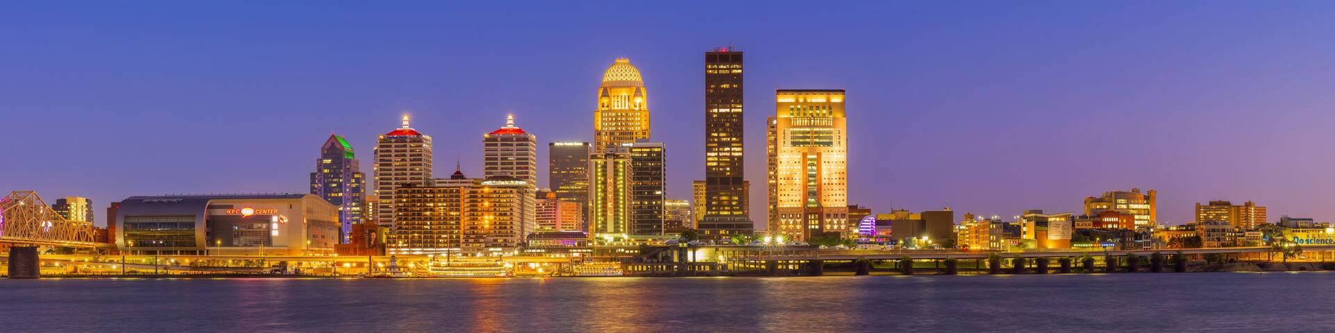 A city skyline at dusk with illuminated buildings reflecting on the water.