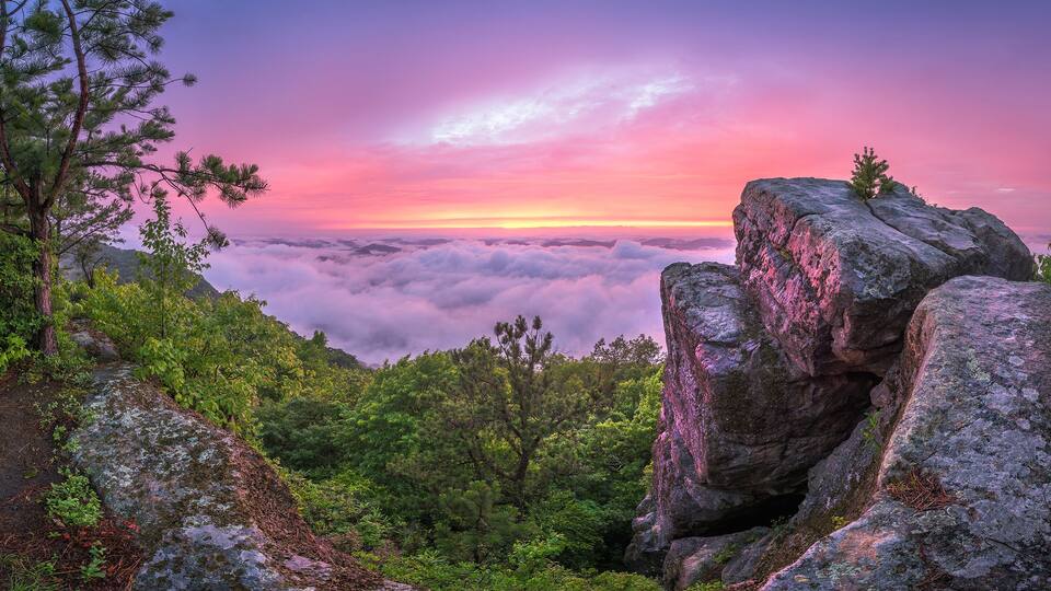 A mountainous landscape with a view of the clouds below and a sunset in the background.