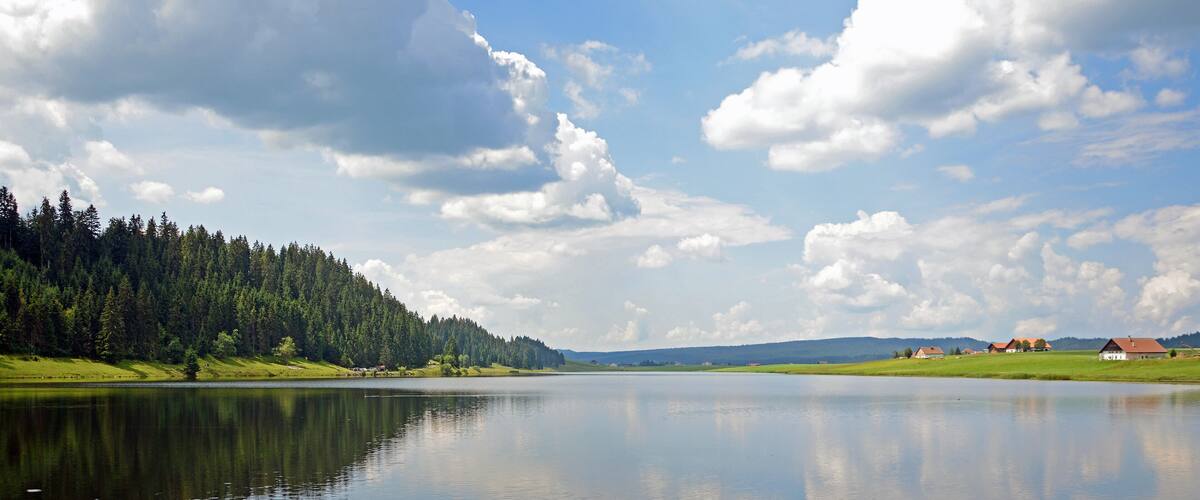 A calm lake with a forested hillside and a few houses in the distance.