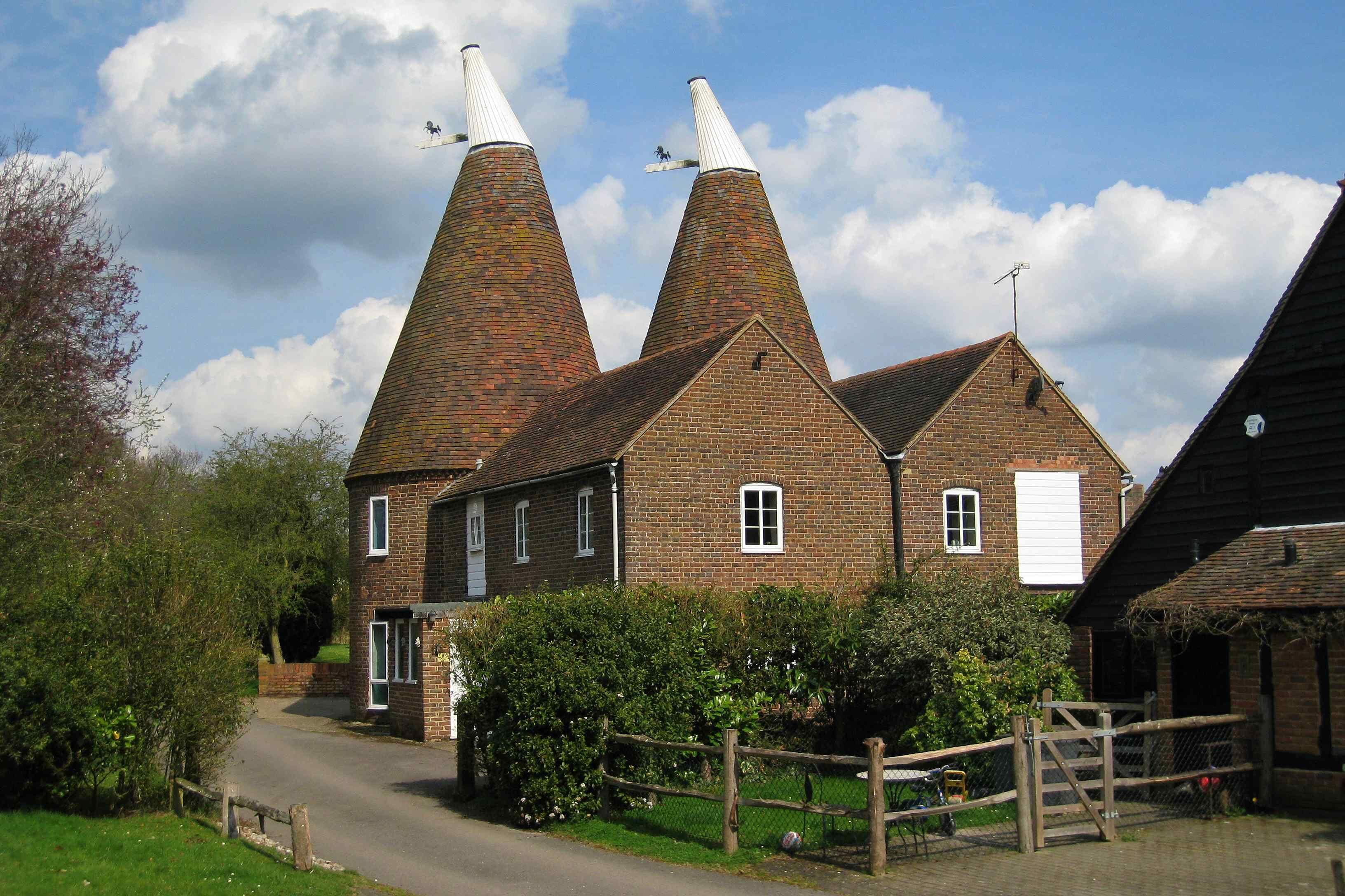 A traditional brick building with two conical roofs, surrounded by greenery and a wooden fence.