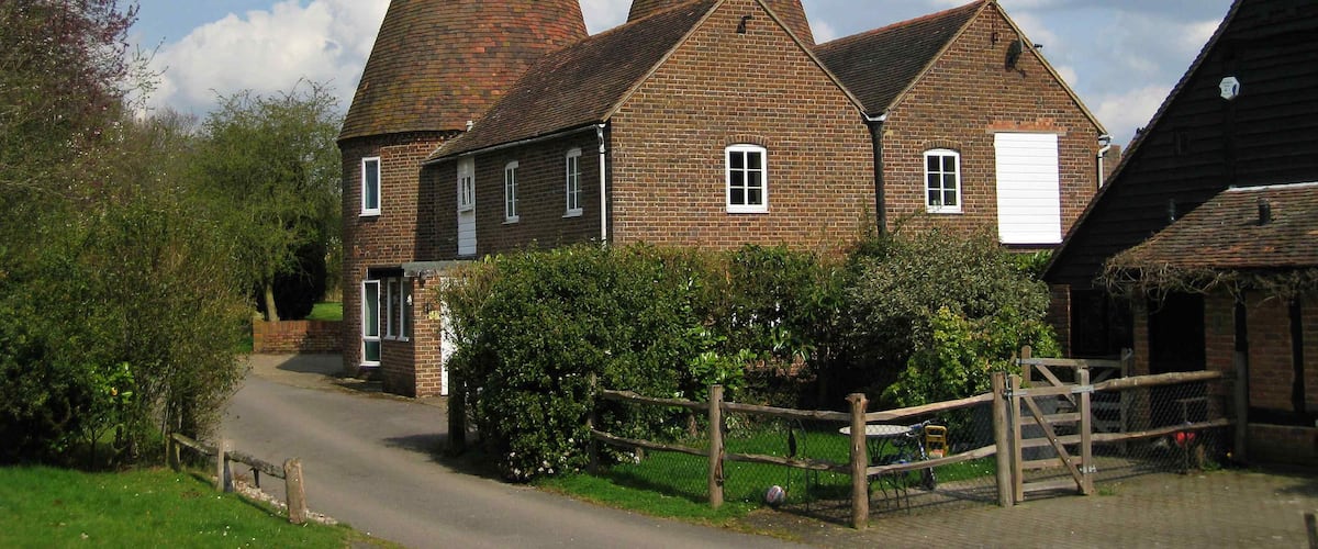 A traditional brick building with two conical roofs, surrounded by greenery and a wooden fence.