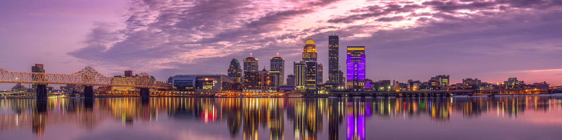 A city skyline at dusk with illuminated buildings reflecting on calm water.