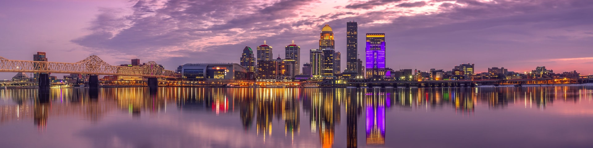 Horizonte urbano al anochecer con edificios iluminados reflejándose en agua tranquila.