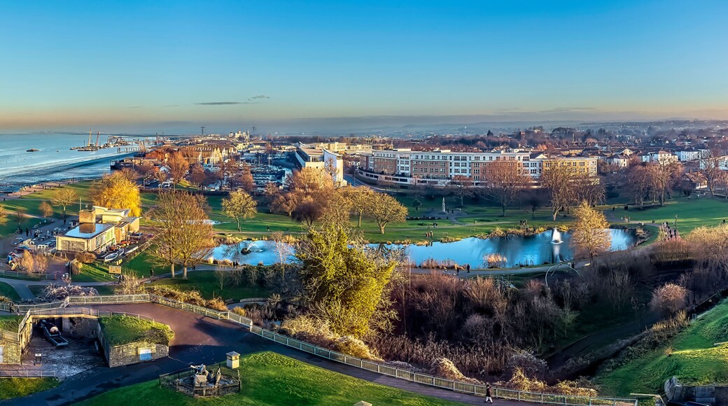 A coastal cityscape with a river, boats, and a park with a fountain.