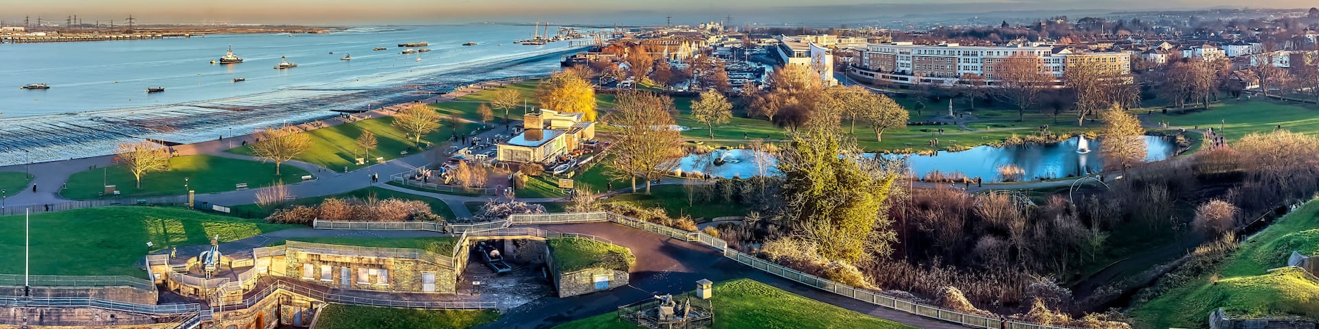 A coastal cityscape with a river, boats, and a park with a fountain.