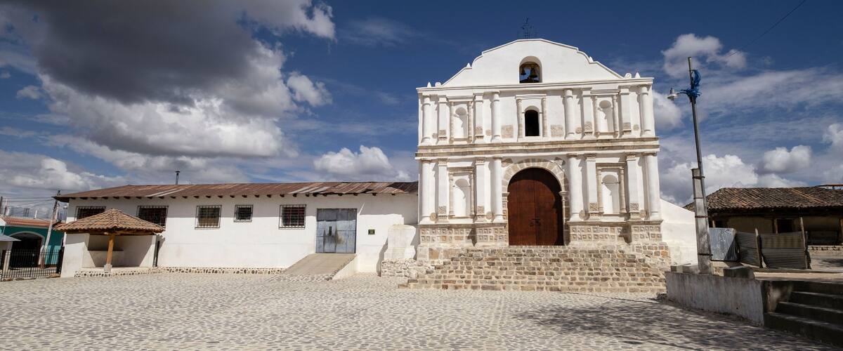 A historic white church with a wooden door and a bell tower, set against a clear blue sky with scattered clouds.