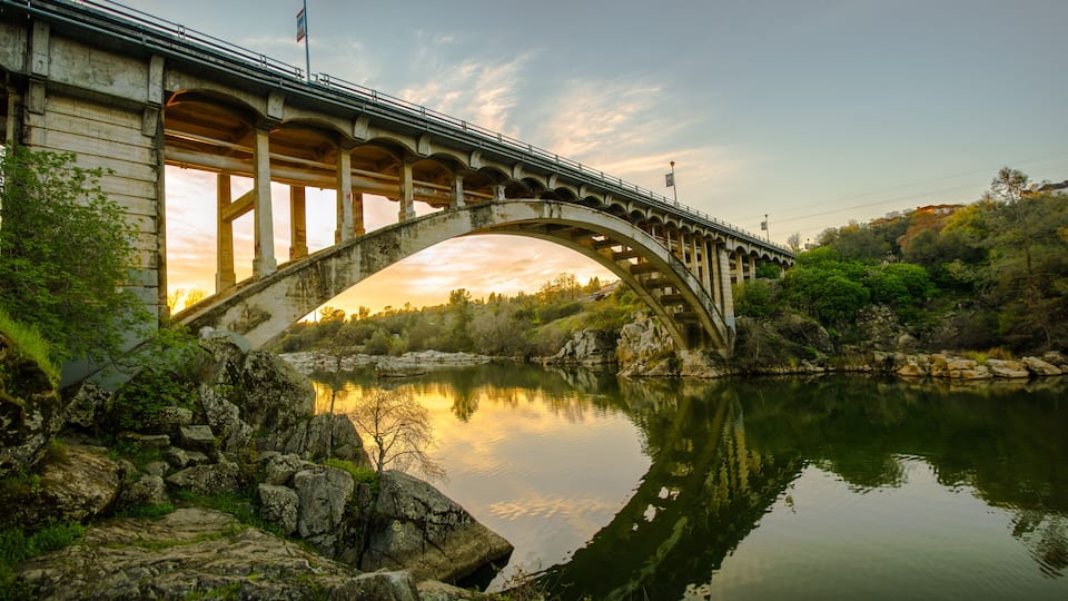 A stone bridge arching over a calm river, surrounded by greenery and rocks.