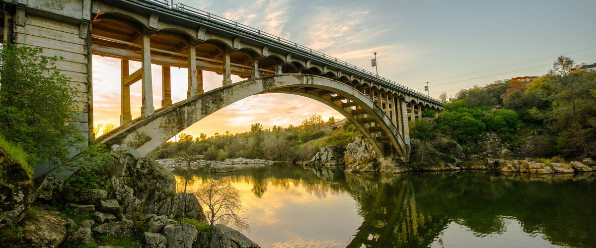 A stone bridge arching over a calm river, surrounded by greenery and rocks.