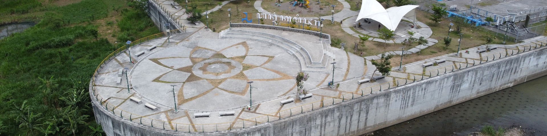 Una gran represa de concreto con una plaza circular que presenta un diseño en forma de estrella, rodeada de áreas verdes y un pequeño parque infantil.