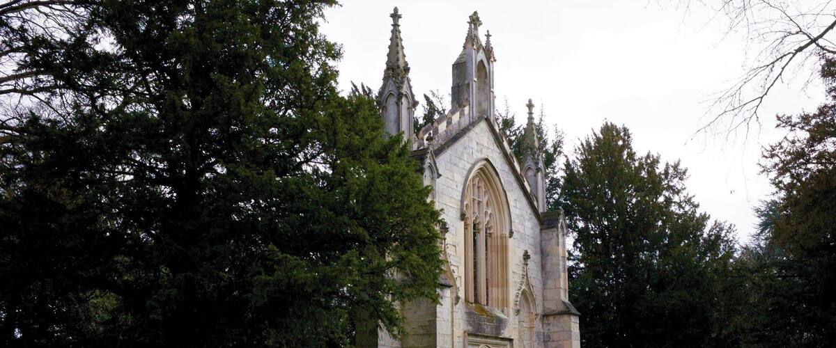 A historic stone church with a tall tower and pointed arches, surrounded by trees and a grassy area.