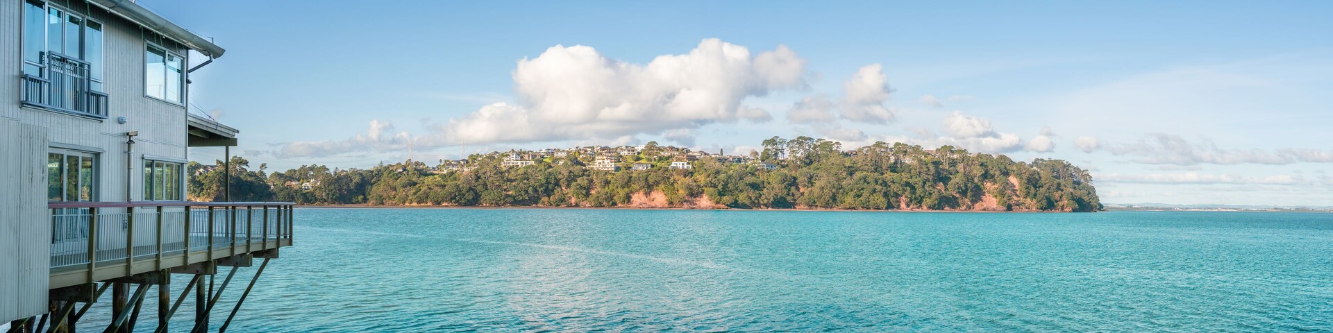 A waterfront property with a wooden deck overlooking turquoise waters, a distant forested island, and a clear sky.