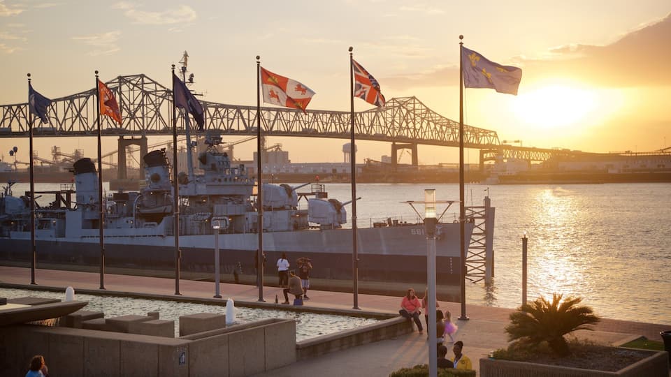 A naval ship docked at a port with multiple flags, a bridge, and a sunset over the water.