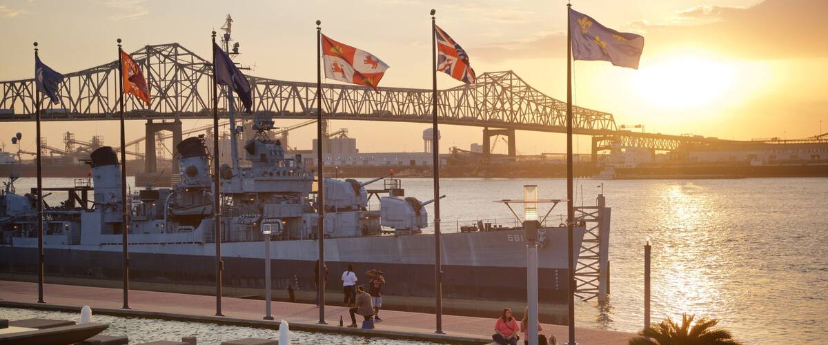A naval ship docked at a port with multiple flags, a bridge, and a sunset over the water.