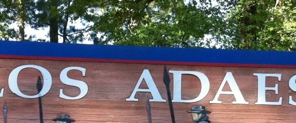 A wooden sign with 'LOS ADAES State Historic Site' and a depiction of soldiers on horseback.