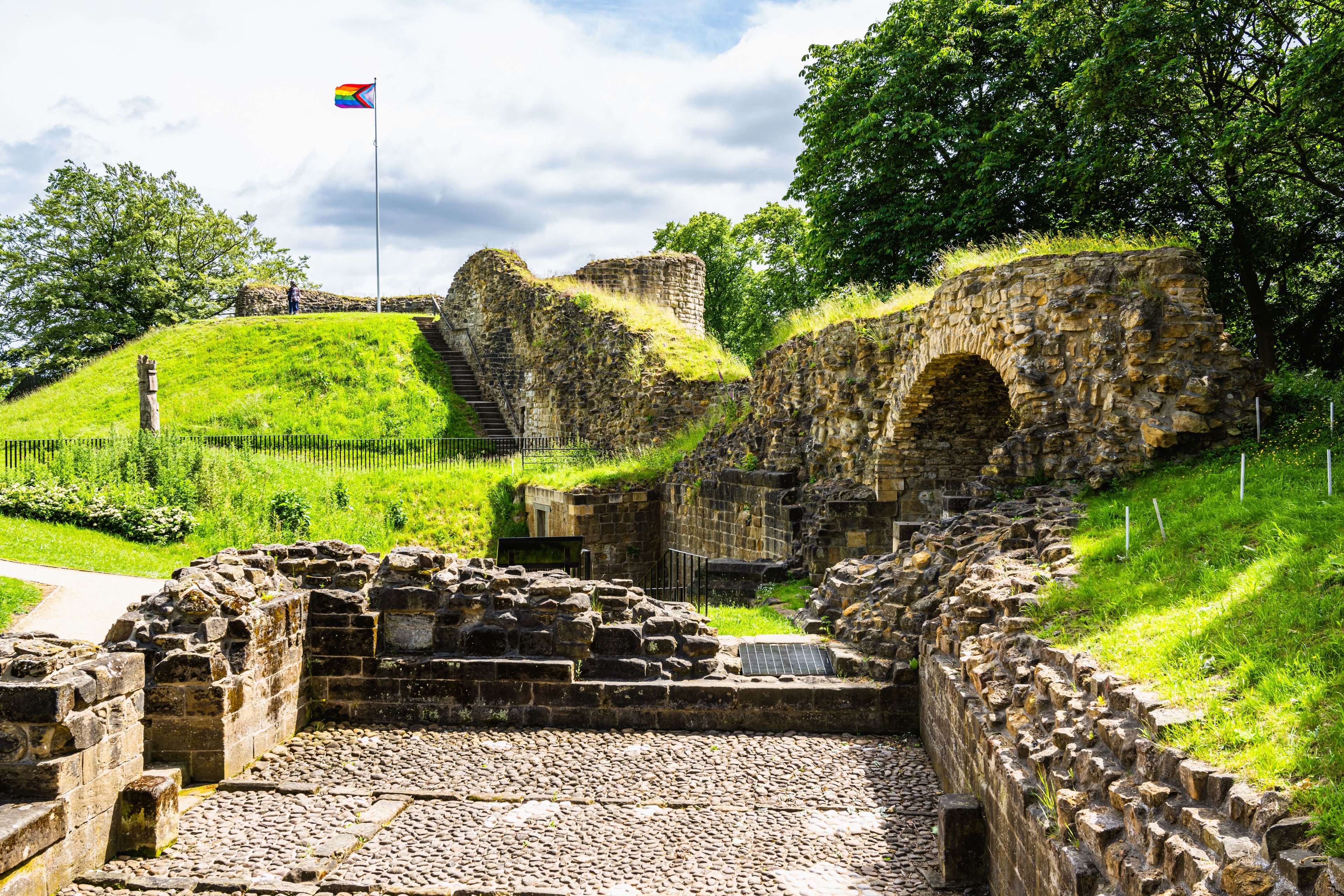 An ancient stone structure with a flag, surrounded by lush greenery and a clear sky.