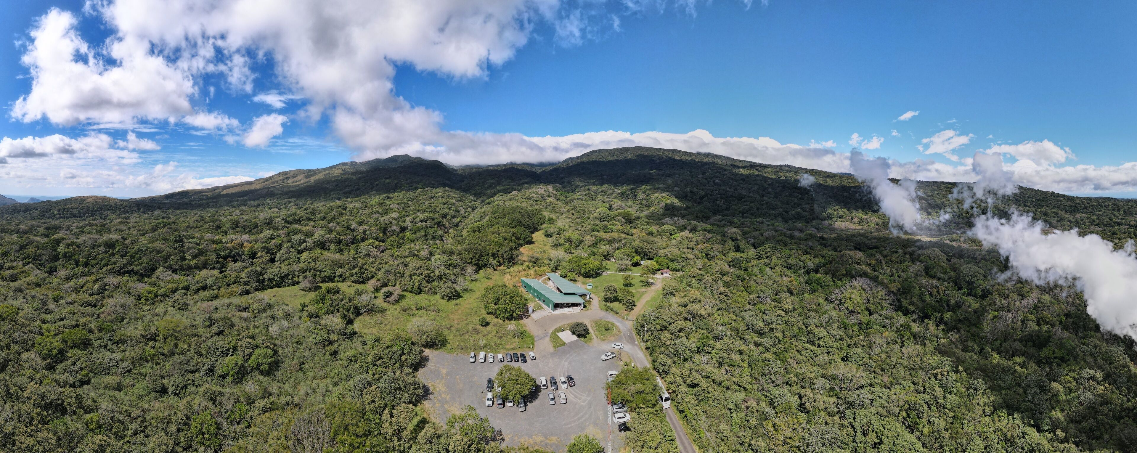 An aerial view of a mountainous area with a parking lot, a building with a green roof, and steam rising from the mountain.