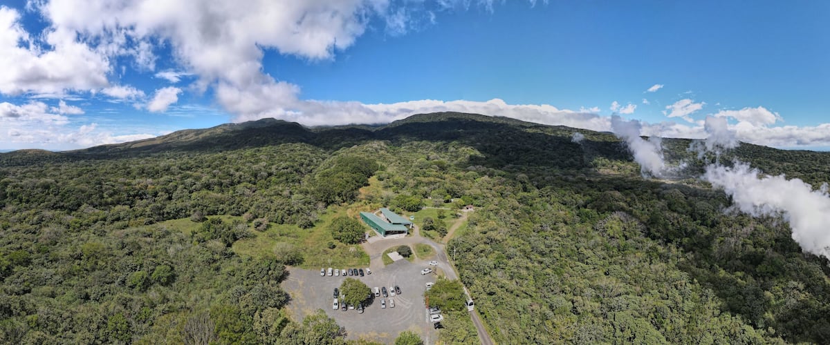 An aerial view of a mountainous area with a parking lot, a building with a green roof, and steam rising from the mountain.