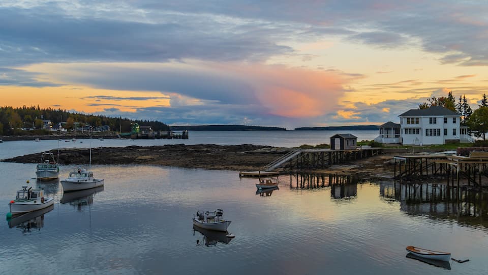 A coastal scene with boats, a dock, a white house, and a forested island in the background.