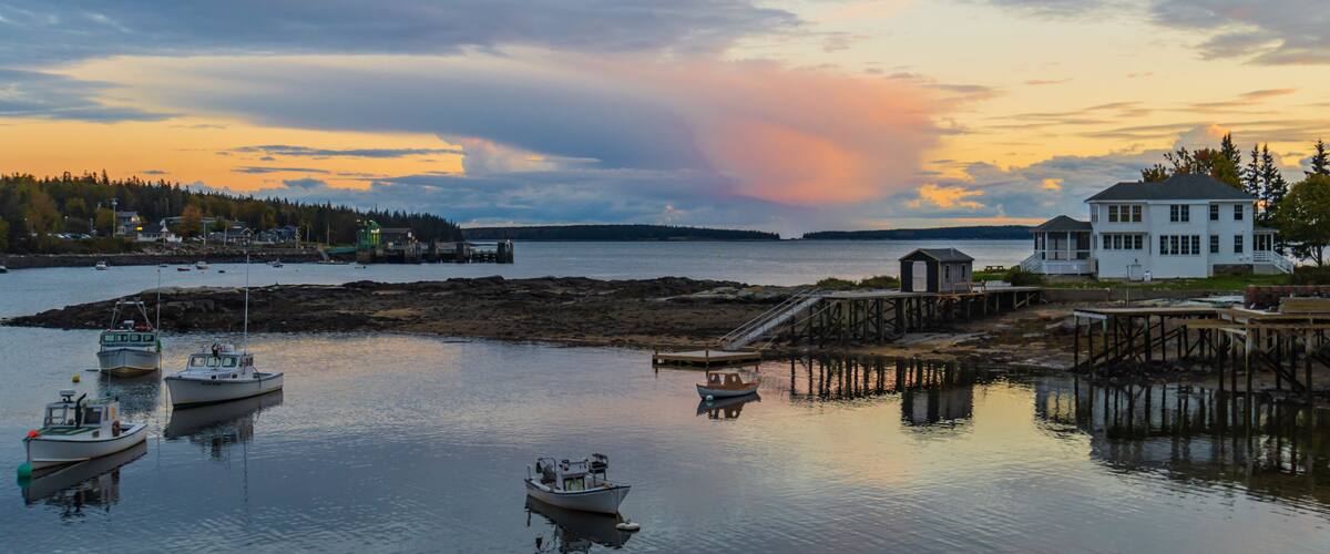 A coastal scene with boats, a dock, a white house, and a forested island in the background.