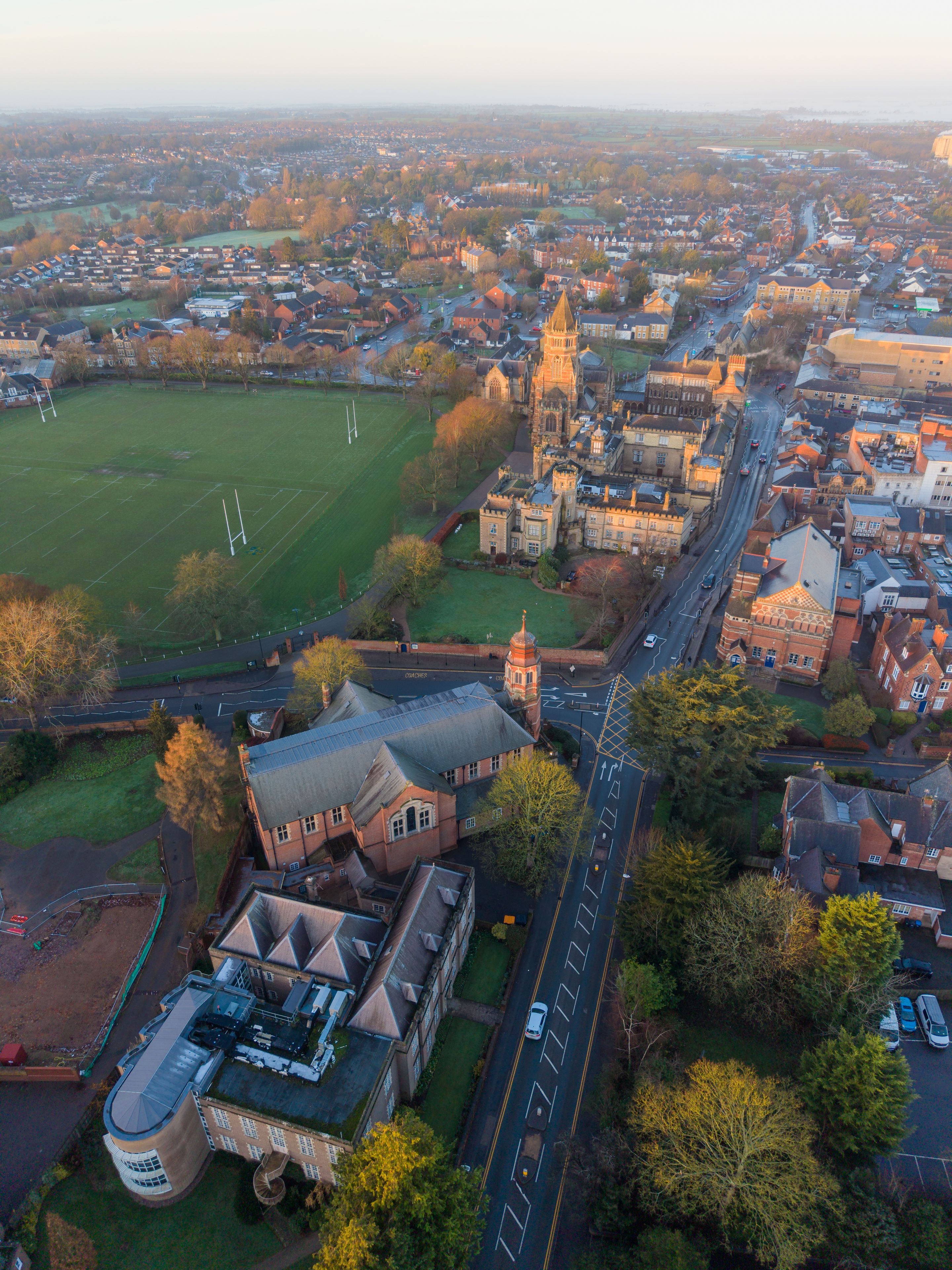 Aerial view of golden sunlight bathing Rugby School's historic buildings and grounds with the town sprawling beyond, a blend of old and new, Rugby, England, United Kingdom.