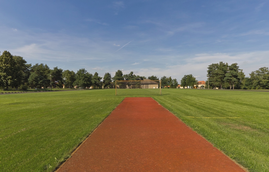Un campo deportivo con una pista roja y un poste de portería, rodeado de árboles y edificios.