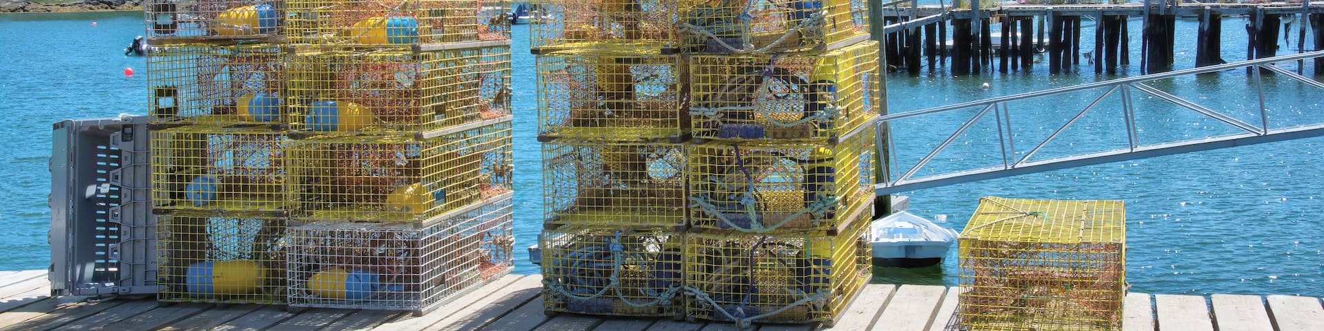 Stacks of yellow lobster traps on a wooden dock by a body of water with a pier in the background.