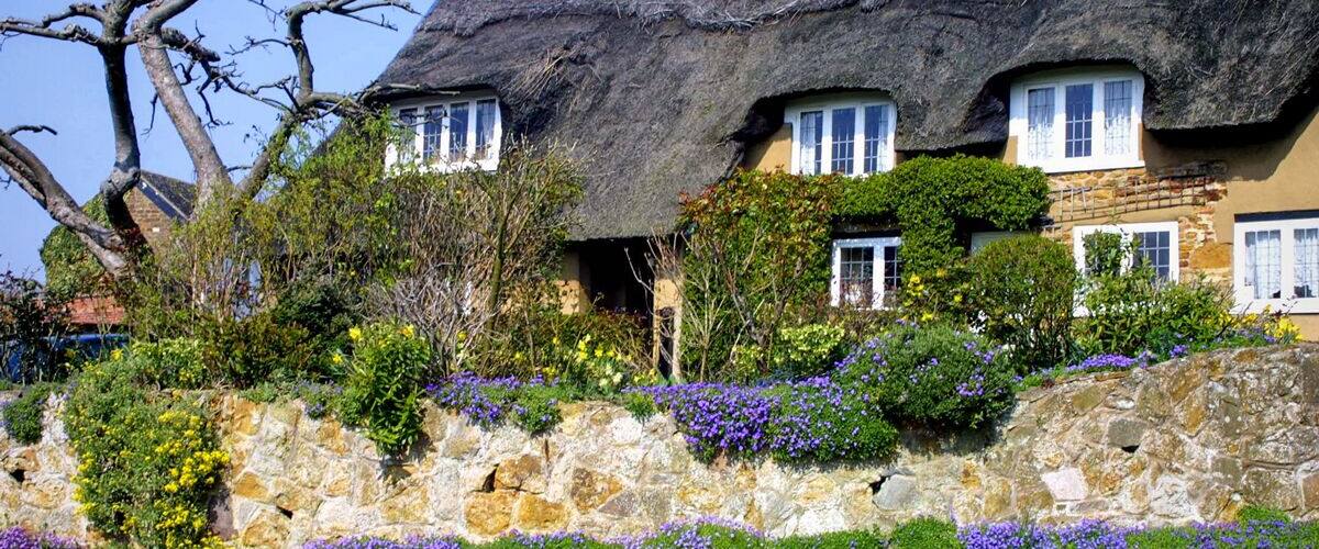 A thatched-roof house with a stone wall and flowering plants.