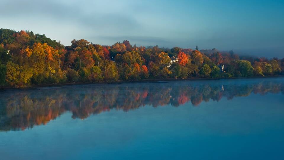 A calm lake with autumn foliage reflecting on the water, surrounded by trees and a distant house.