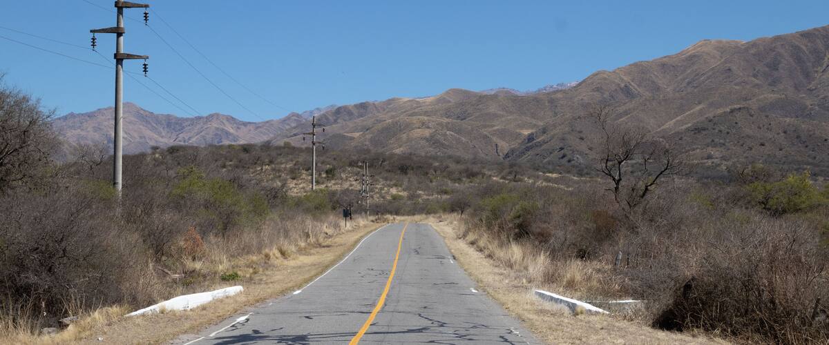 A straight road with a yellow center line, flanked by dry grass and shrubs, leading towards a mountain range under a clear blue sky.