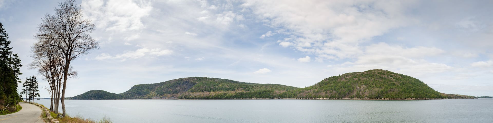 A calm lake with two hills in the background and a paved path on the left.