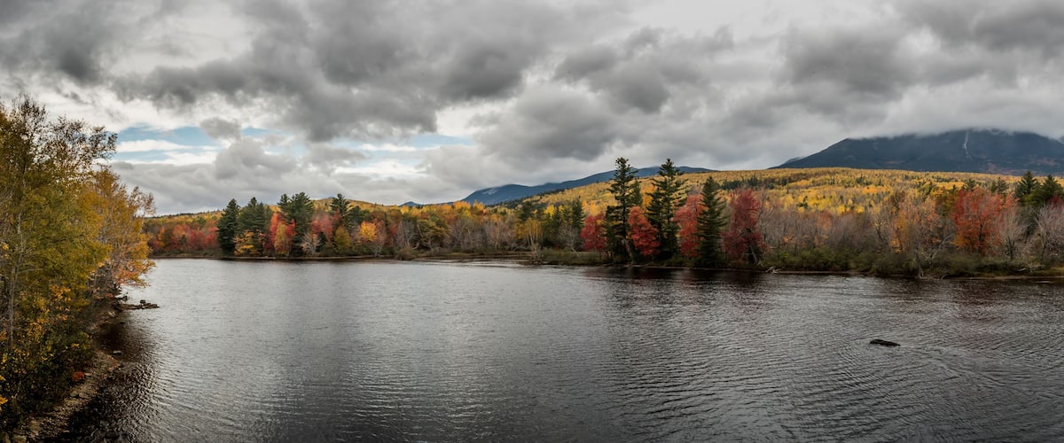 A river with autumn foliage, a mountain range, and a cloudy sky.