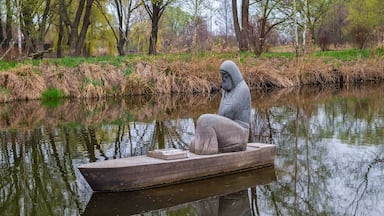 Estátua de uma figura encapuzada em um barco, em um rio calmo, cercada por árvores e vegetação.