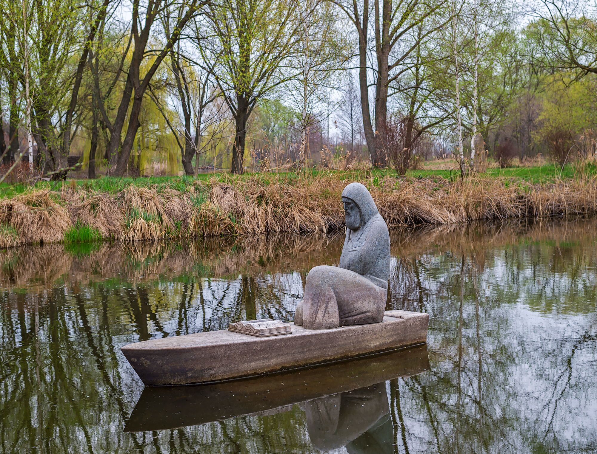 A statue of a hooded figure in a boat on a calm river, surrounded by trees and greenery.