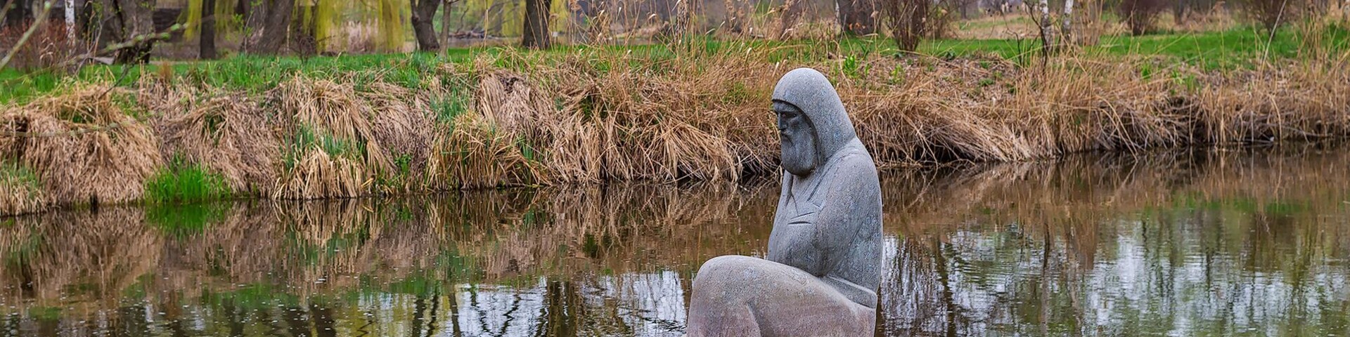 A statue of a hooded figure in a boat on a calm river, surrounded by trees and greenery.