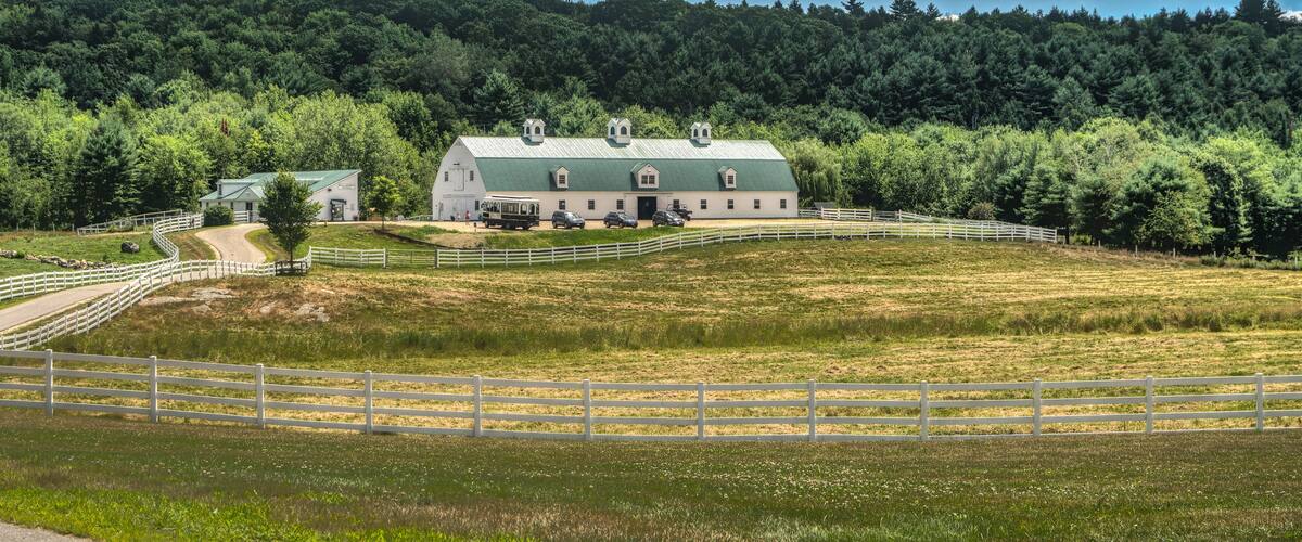 A barn with a green roof surrounded by a white fence, set against a backdrop of a forested hill and a clear blue sky.