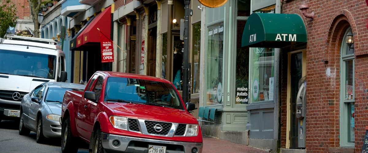 A red Nissan truck parked on a city street, a green ATM, and a building with a sign that reads "House of Jerky Open".