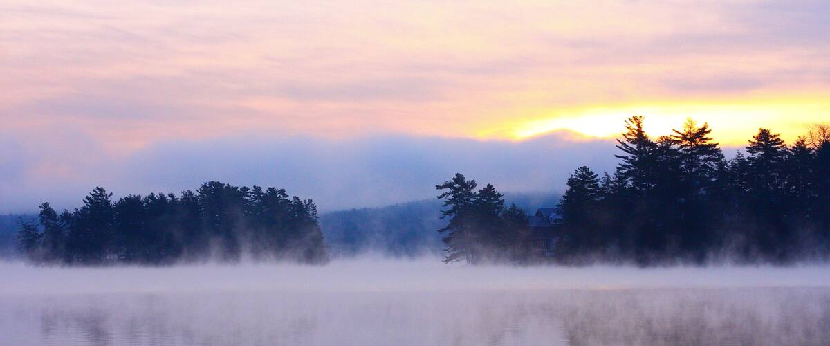 A misty lake with reflections of trees and a sunrise.