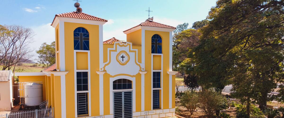 A yellow church with a cross on the facade, surrounded by trees and a clear sky.