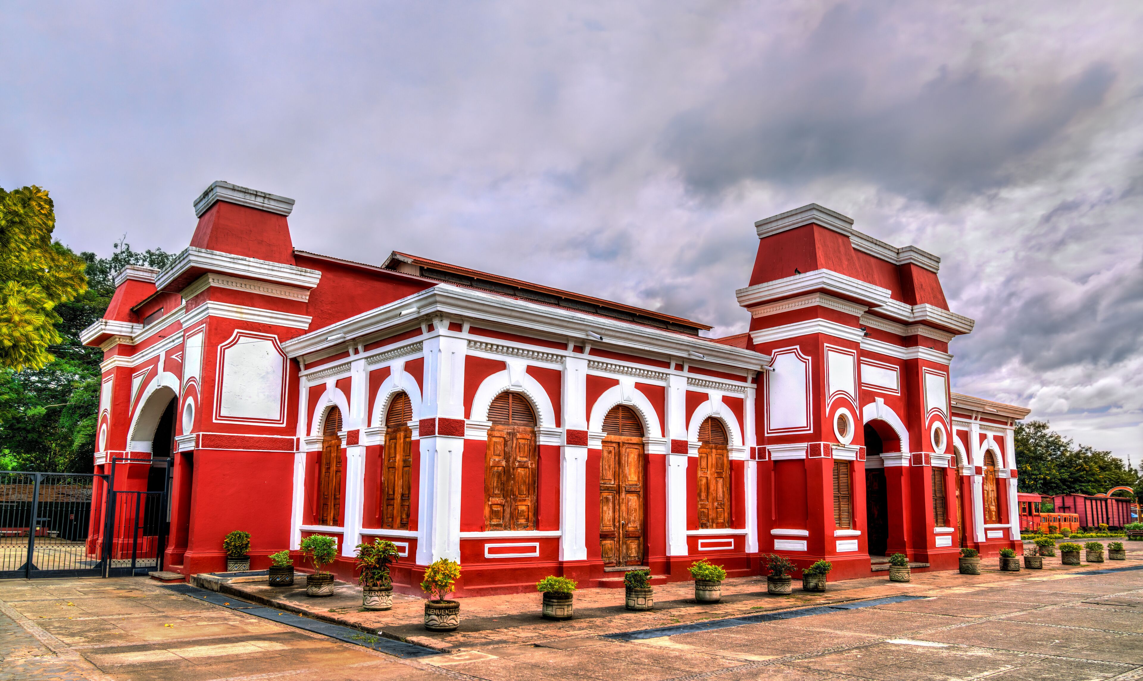 A red and white colonial-style building with arched windows and doors, surrounded by potted plants.