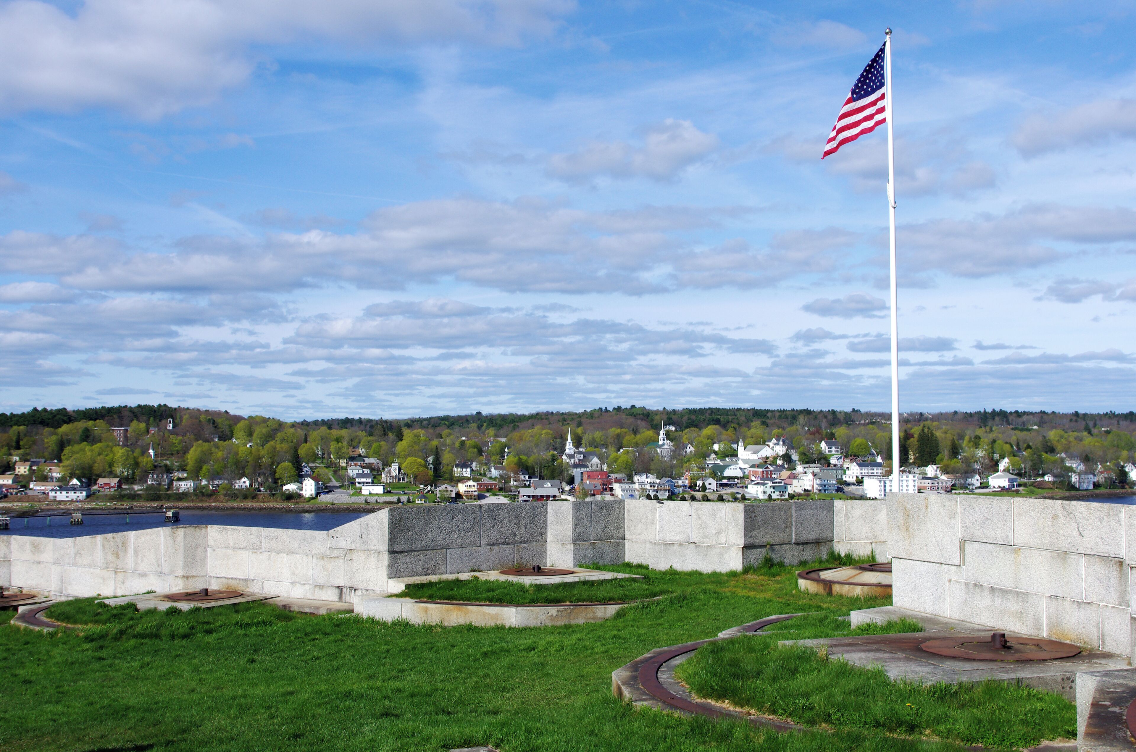 A coastal town with a prominent American flag, a river, and a distant hill with buildings.