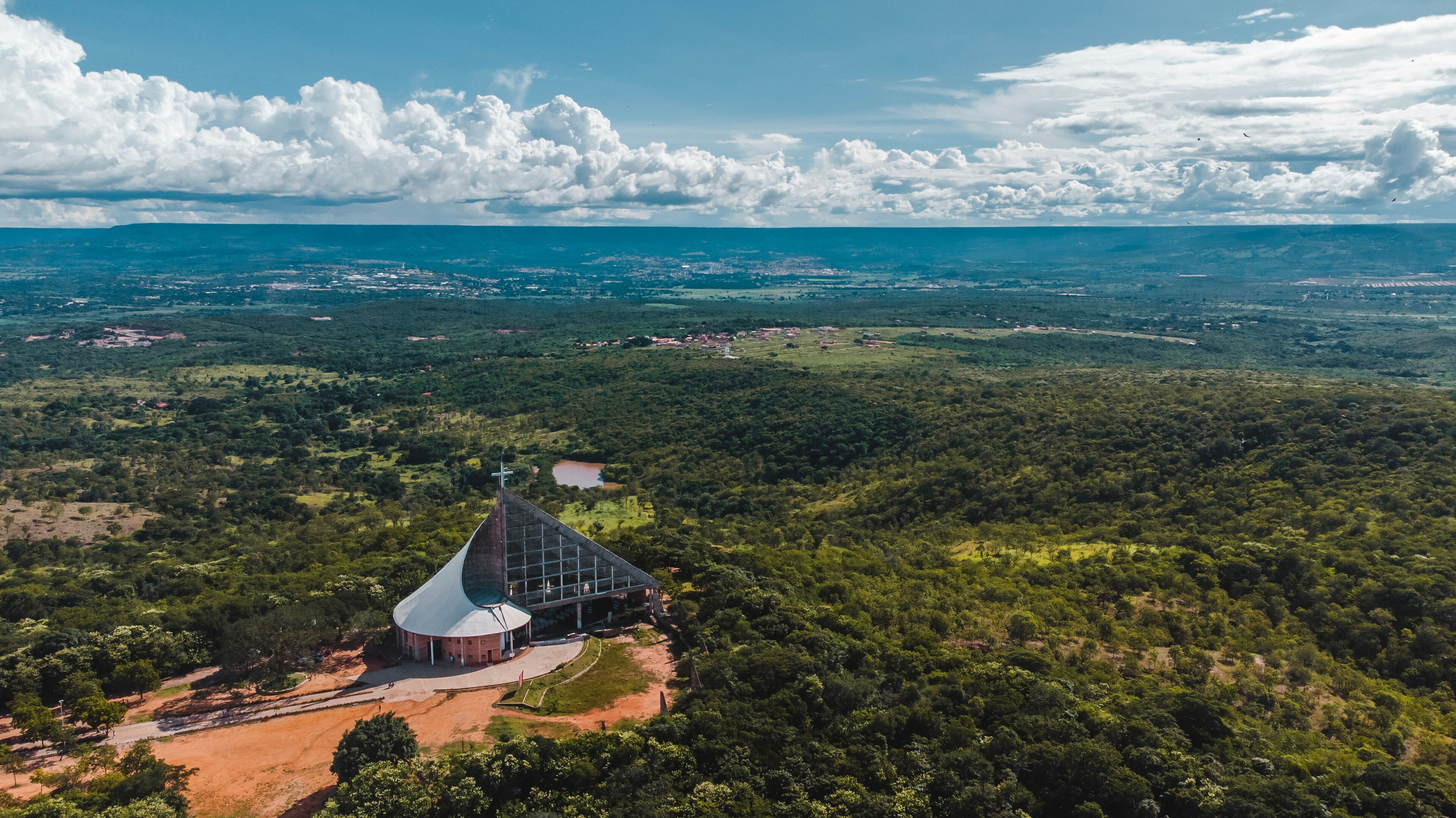 A church with a distinctive roof and a cross atop, surrounded by lush greenery and a vast landscape.